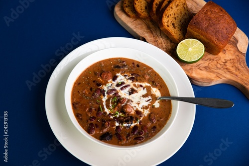 Bean soup with sliced sausages, dried plums and cream in white plate,bread on board, all on blue background. photo