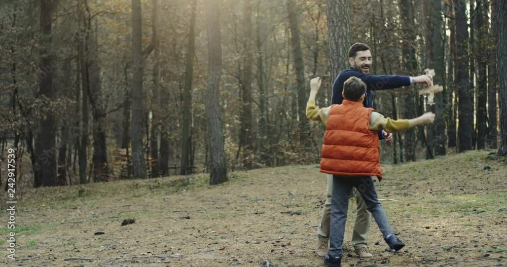 Cheerful Caucasian dad playing with a wooden plane and his small son trying to reach it and take it from father in the wood. Outside.