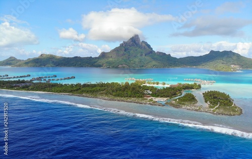 Aerial panoramic landscape view of the island of Bora Bora in French Polynesia with the Mont Otemanu mountain surrounded by a turquoise lagoon, motu atolls, reef barrier, and the South Pacific Ocean
