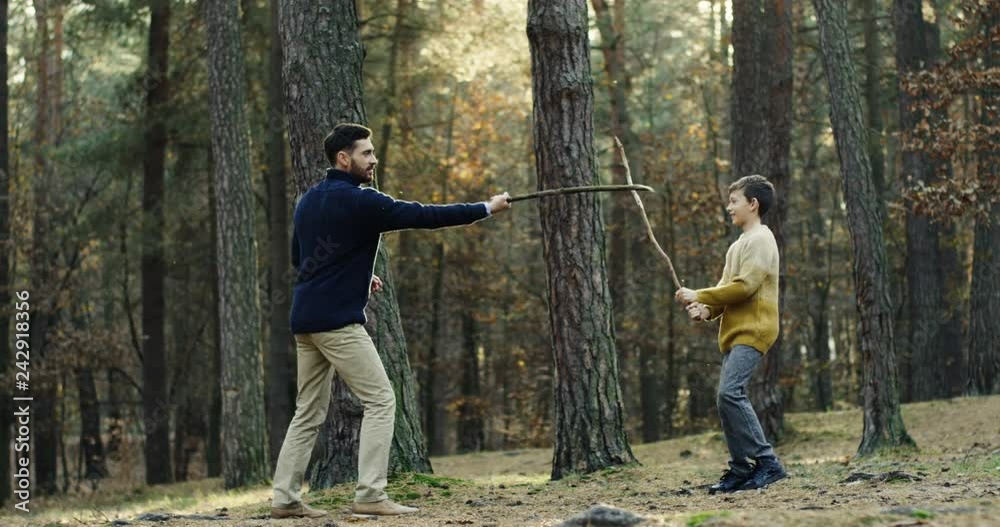 Handsome dad teaching his son to fight on swords while they playing with sticks in the forest. Outside. Handsome dad teaching his son to fight on swords while they playing with sticks in the forest. Outside.
