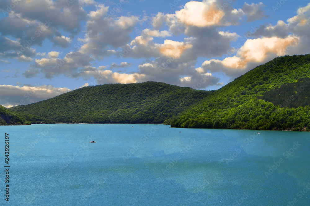 Mountains with green forest on a lake shore on the blue sky with clouds background