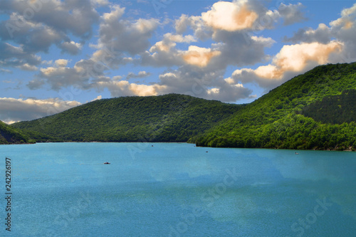 Mountains with green forest on a lake shore on the blue sky with clouds background