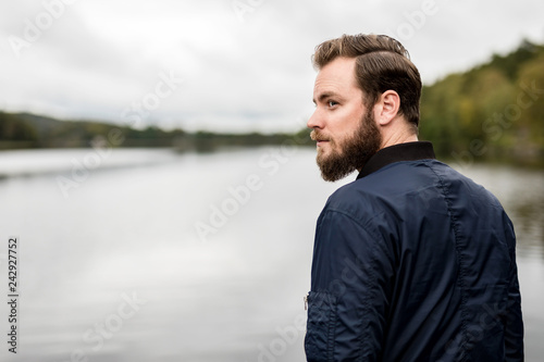 A pensive bearded man wearing a blue jacket and jeans, standing outside with a big lake with surrounding woods.
