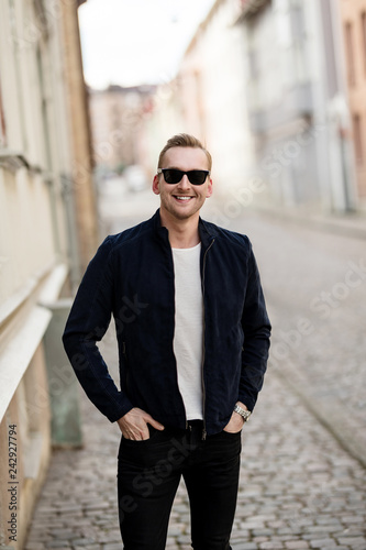 Confident fashionable man standing near a wall in an alley wearing a blue leather jacket and sunglasses smiling towards camera.