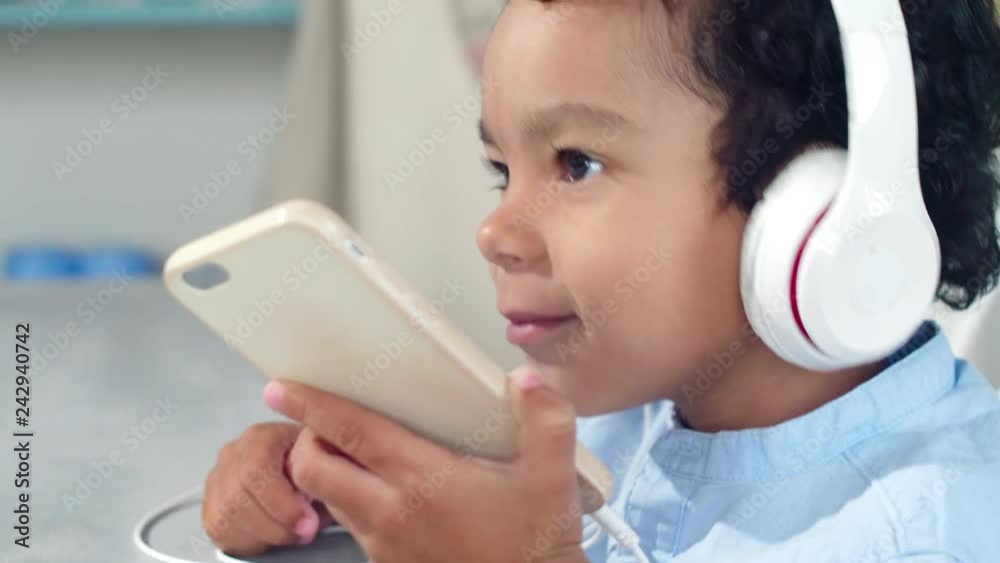 Tilt up of joyous little boy of African ethnicity holding smartphone while listening to music in headphones, smiling and bobbing his head