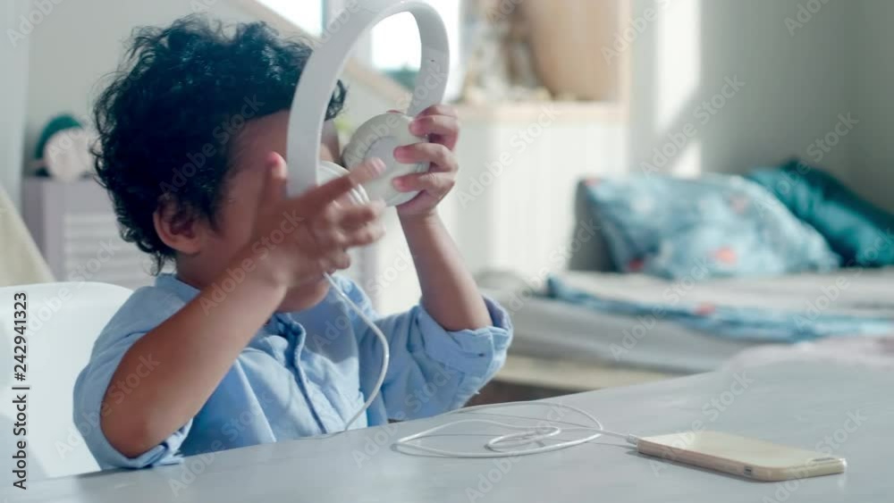 Happy little african boy putting on headphones and start bobbing his head while listening to music on smartphone at home