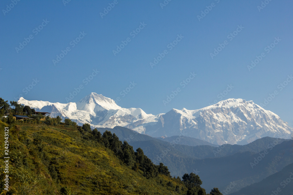 green slope of a hill with houses on the background of the snowy mountain ridge of Annapurna under a clear blue sky