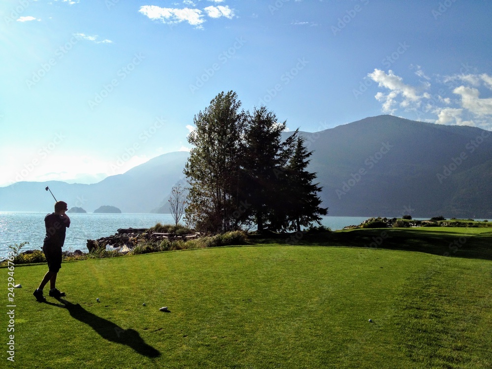 A man playing golf along the ocean in howe sound, British Columbia, Canada.  It is a beautiful sunny day.  He is playing a par 3 and is following through after hitting the ball.