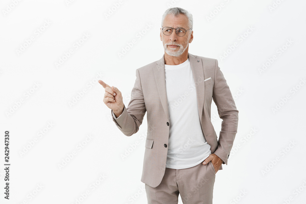 Unretouched shot of good-looking intelligent and elegant old man with grey beard and hair holding hand in pocket pointing and looking at upper left corner with questioned uncertain and disliking look Unretouched shot of good-looking intelligent and elegant old man with grey beard and hair holding hand in pocket pointing and looking at upper left corner with questioned uncertain and disliking look
