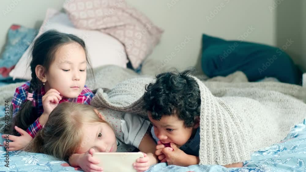 Cute little Asian and Caucasian girls and African boy lying together on bed under cozy blanket and watching cartoon on mobile phone Cute little Asian and Caucasian girls and African boy lying together on bed under cozy blanket and watching cartoon on mobile phone