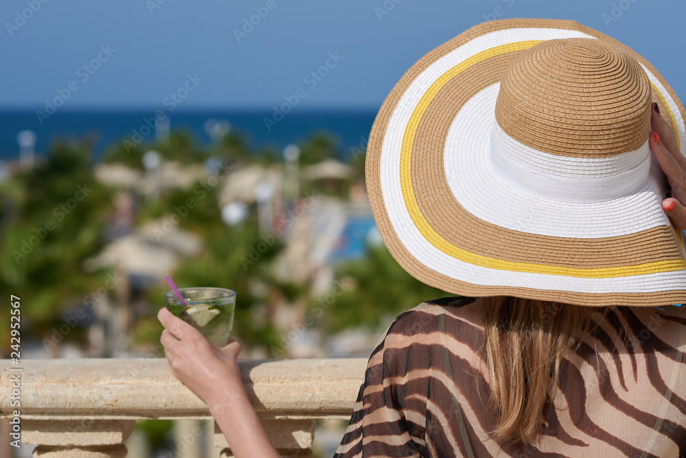 Beautiful European girl in a bonnet hat is relaxing on resort balcony with glass of mojito against sea.