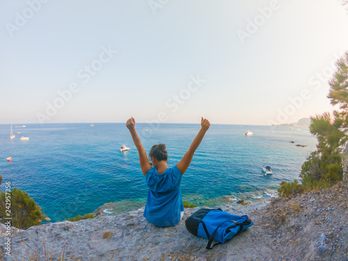 Young woman with backpack sitting on a cliff looking at the ocean spreading arms. Ponza Island coast, Italy.