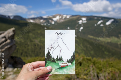 The travel postcard in the hand with background of mountains, adventures in the mountains, enjoy the moment
