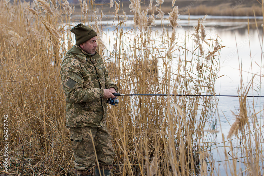 Fishing in the reeds for spinning on quiet water. A man in camouflage clothes in cool weather is fishing on the river bank. Fishing in the reeds for spinning on quiet water. A man in camouflage clothes in cool weather is fishing on the river bank.