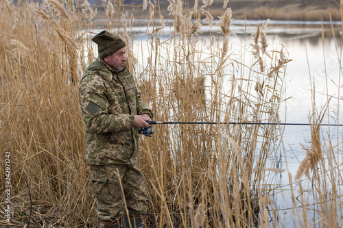 Fishing in the reeds for spinning on quiet water. A man in camouflage clothes in cool weather is fishing on the river bank. © Mountains Hunter