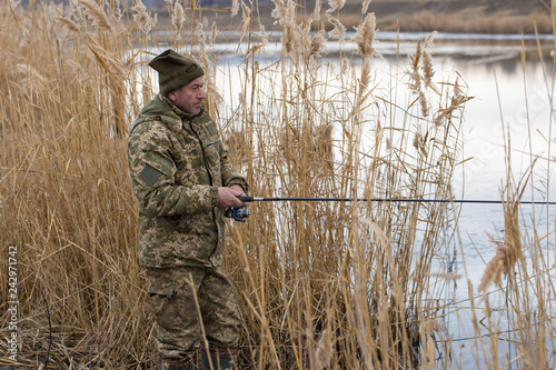 Fishing in the reeds for spinning on quiet water. A man in camouflage clothes in cool weather is fishing on the river bank.