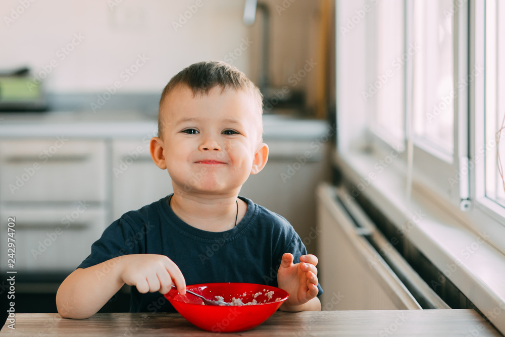 A child in the kitchen eating their own oatmeal with a red plate A child in the kitchen eating their own oatmeal with a red plate