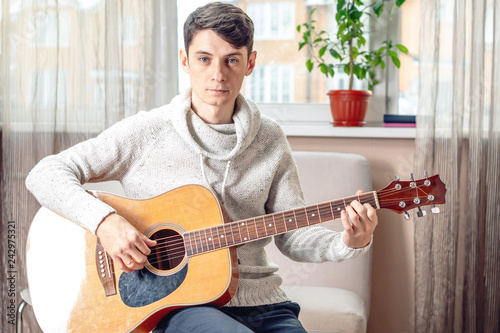 Young attractive male musician sitting on a chair playing acoustic guitar in room. Concept of music as a hobby