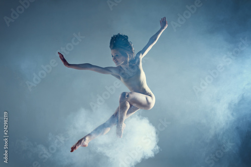 Dancing in cloud concept. Muscle brunette beauty female girl adult woman dancer athlete in dust / fog. Girl wearing dance bodysuit jumping in mid air, performance on isolated grey / black background