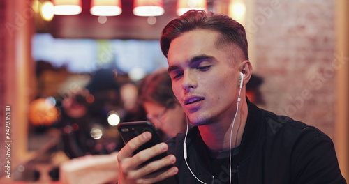 Handsome young man uses headphones and his smartphone for video call. He sits at the bar near the neon signs. Cheerful mood, modern communication, technologies, devices