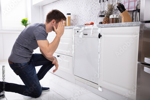 Upset Man Sitting In Front Of Damaged Dishwasher