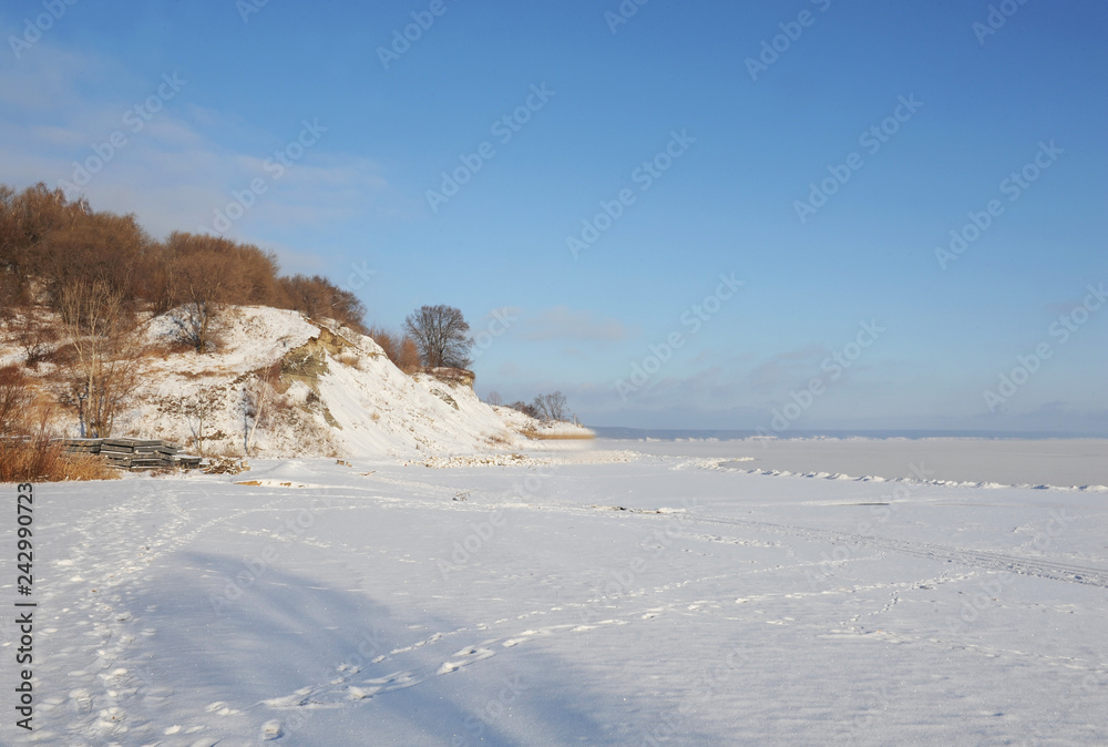Winter snowy coastal landscape on a sunny day. Frozen river. Clear sky. Open space. Hill with trees