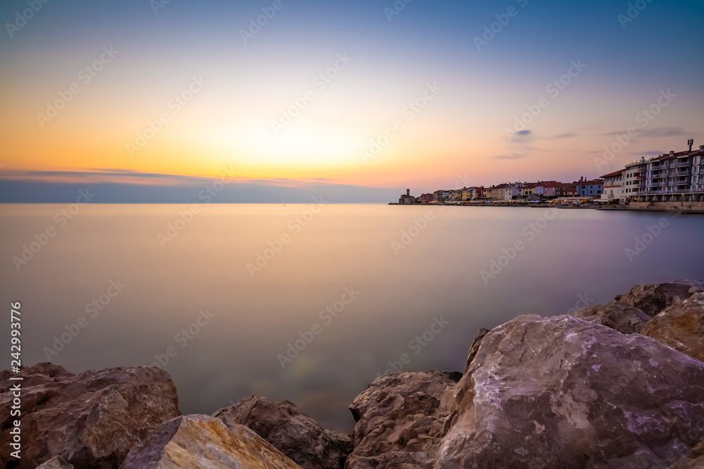 Sunset above the adriatic sea in Piran city, Slovenia. Old ancient and mediaveal city, view from coast and harbor. Old stones in foreground, beautiful clouds on sky, romantic sunset above city.
