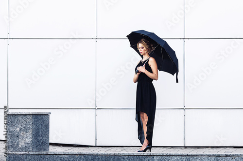 elegant young girl in a black dress and with an umbrella posing against a gray wall.