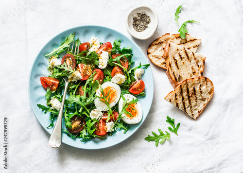 Breakfast salad - arugula, cherry tomatoes, mozzarella and boiled egg with olive oil, mustard, lemon dressing on a light background, top view