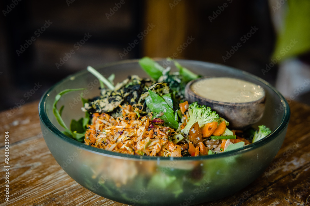 Vegan salad with various veggies and greens, and dressing in a glass bowl on a wooden table. Closeup, food blog, healthy eatingrecipe