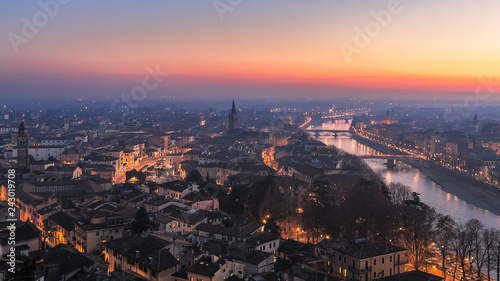 Panoramic view of the city of Verona with night city illumination and the Adige river with bridges covered with the evening mist colored by the setting sun. Sunset in Verona. Winter time. Italy.