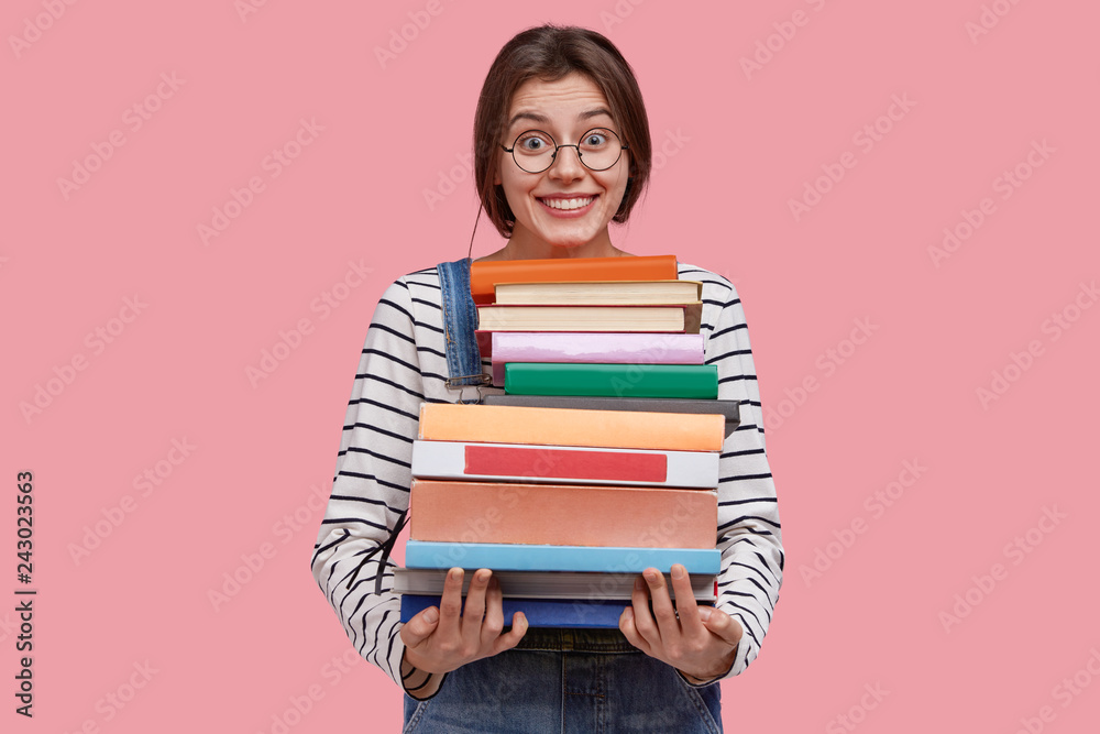 Delighted young woman carries pile of textbooks, smiles broadly, learns useful information from encyclopedia, has dark hair, dressed in casual outfit, poses aginst pink background. Education concept Delighted young woman carries pile of textbooks, smiles broadly, learns useful information from encyclopedia, has dark hair, dressed in casual outfit, poses aginst pink background. Education concept