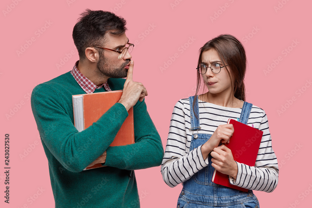 Φωτογραφία Young male teacher or tutor holds red old textbook, shows silence sign, asks trainee not spread rumours, tells secret information, look at each other