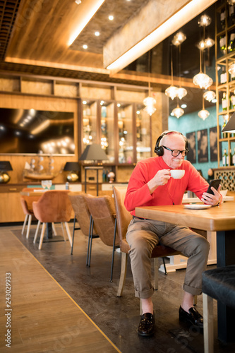Content satisfied handsome elderly man in stylish turtleneck sitting at table in cafe and reading internet news on smartphone while listening to music via headphones and drinking coffee