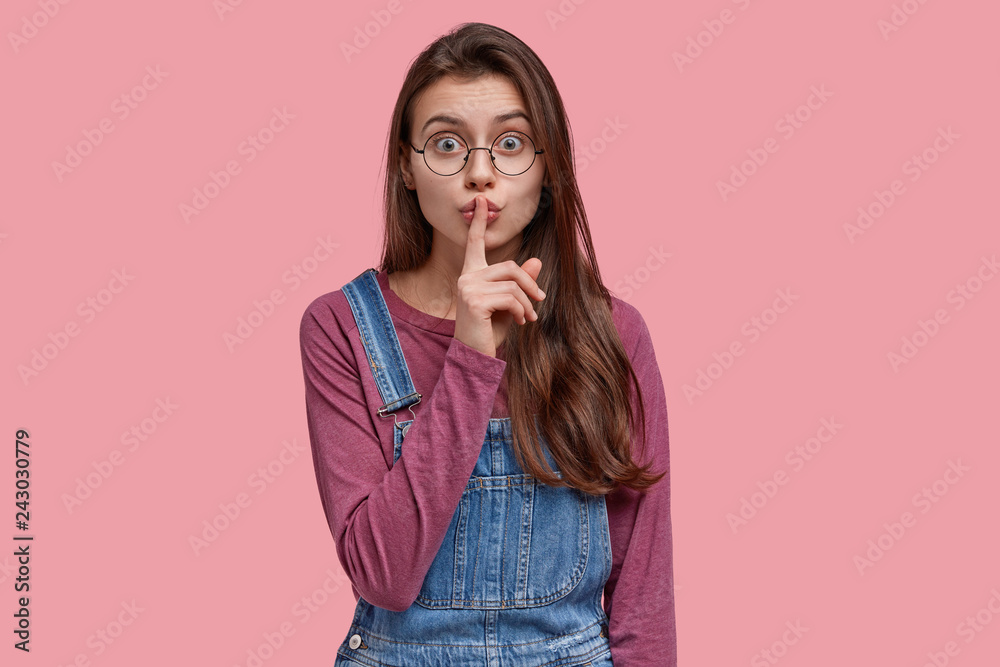 Mysterious beautiful young lady makes shush gesture, wears denim overalls, asks not tell her secret, has optical glasses, isolated over pink background, gossips about latest news with friend Mysterious beautiful young lady makes shush gesture, wears denim overalls, asks not tell her secret, has optical glasses, isolated over pink background, gossips about latest news with friend