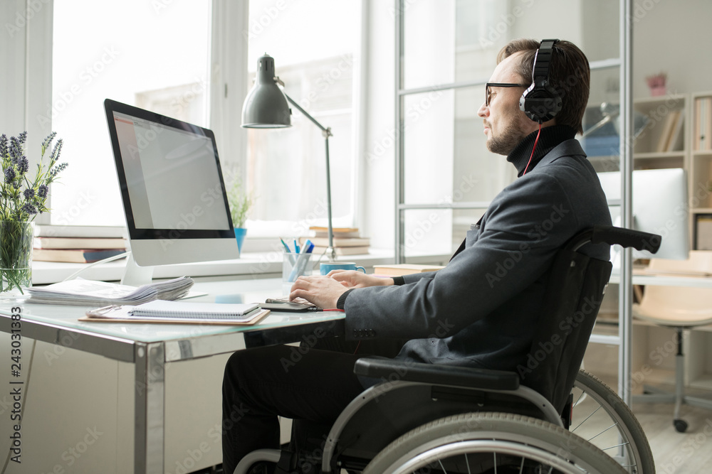 Disabled businessman in wheelchair wearing headphones and concentrated on his work, he typing on keyboard and looking at computer screen - obrazy, fototapety, plakaty 