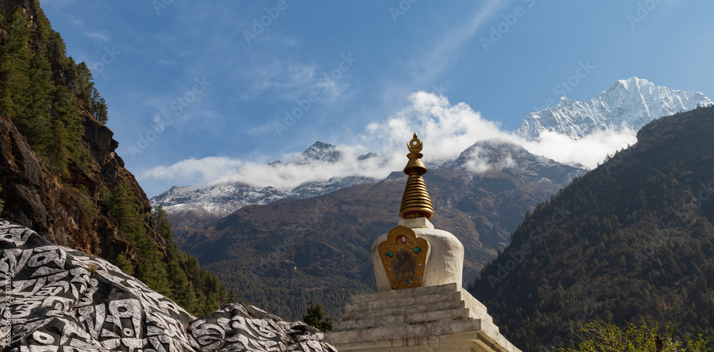 Religious stone and top of temple along the Everest base camp hike in Nepal with mountains and clouds in the background in the valley.