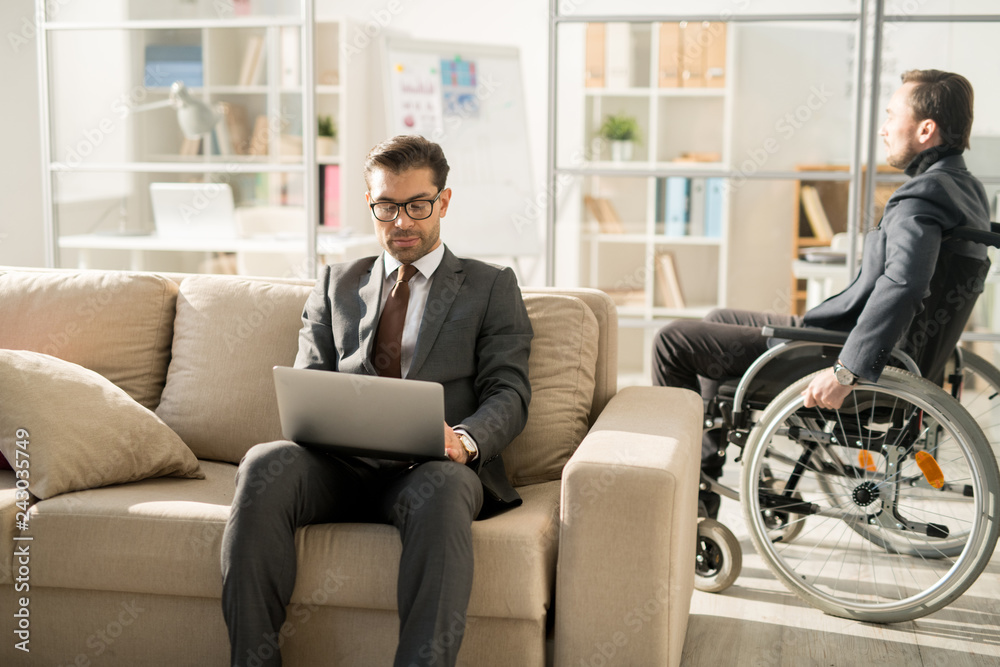 Serious manager in suit sitting on sofa and typing on laptop with disabled businessman moving around the office in wheelchair in the background Serious manager in suit sitting on sofa and typing on laptop with disabled businessman moving around the office in wheelchair in the background