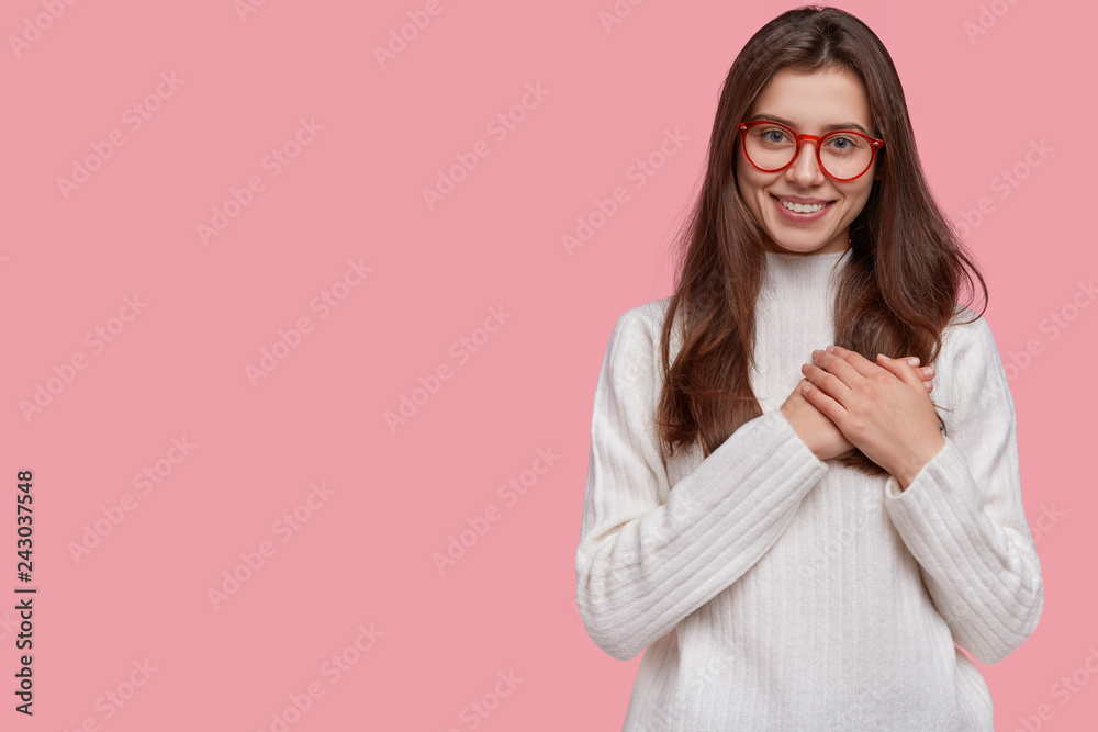 Kind tender girl presses hands to heart in thankful and touched gesture, has pleasant smile, wears white jumper, poses against pink background with copy space for your text. Body language concept