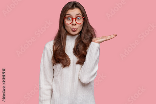 Isolated shot of good looking friendly experienced seller raises hand, demonstrates something against pink background, dressed in casual white sweater, poses against pink background, keeps lips folded