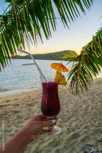 Summer vacation/holiday refreshing exotic cocktail concept. View of purple drink/beverage in glass decorated with plastic straws, umbrellas and piece of pineapple on sandy beach of Fiji Islands. © Dajahof