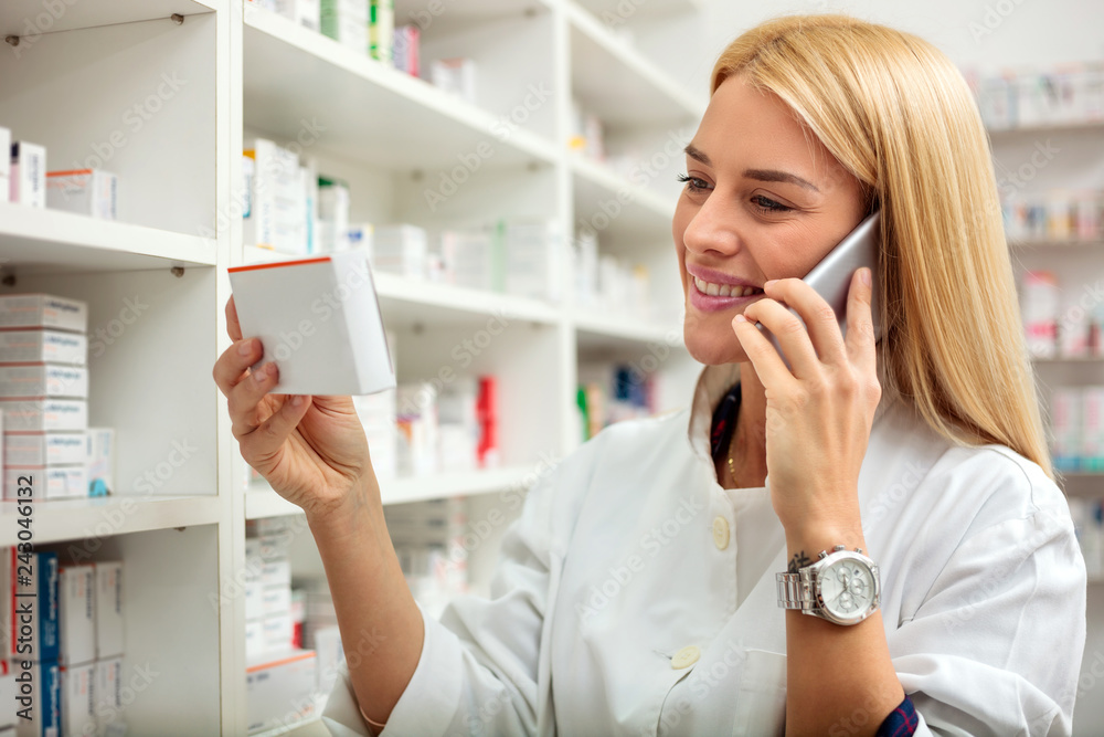 Happy young blond haired female pharmacist checking medication details on a box and talking on a smart phone. Medicine, healthcare and pharmaceutics concept Happy young blond haired female pharmacist checking medication details on a box and talking on a smart phone. Medicine, healthcare and pharmaceutics concept