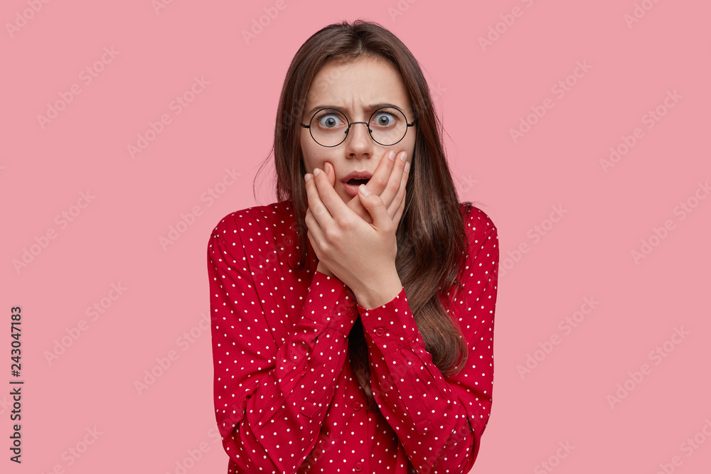 Headshot of frightened lady looks with terrible expression, keeps hands near mouth, hears bad news, has dark hair, wears polka dot red shirt, isolated over pink background. Negative emotions Headshot of frightened lady looks with terrible expression, keeps hands near mouth, hears bad news, has dark hair, wears polka dot red shirt, isolated over pink background. Negative emotions