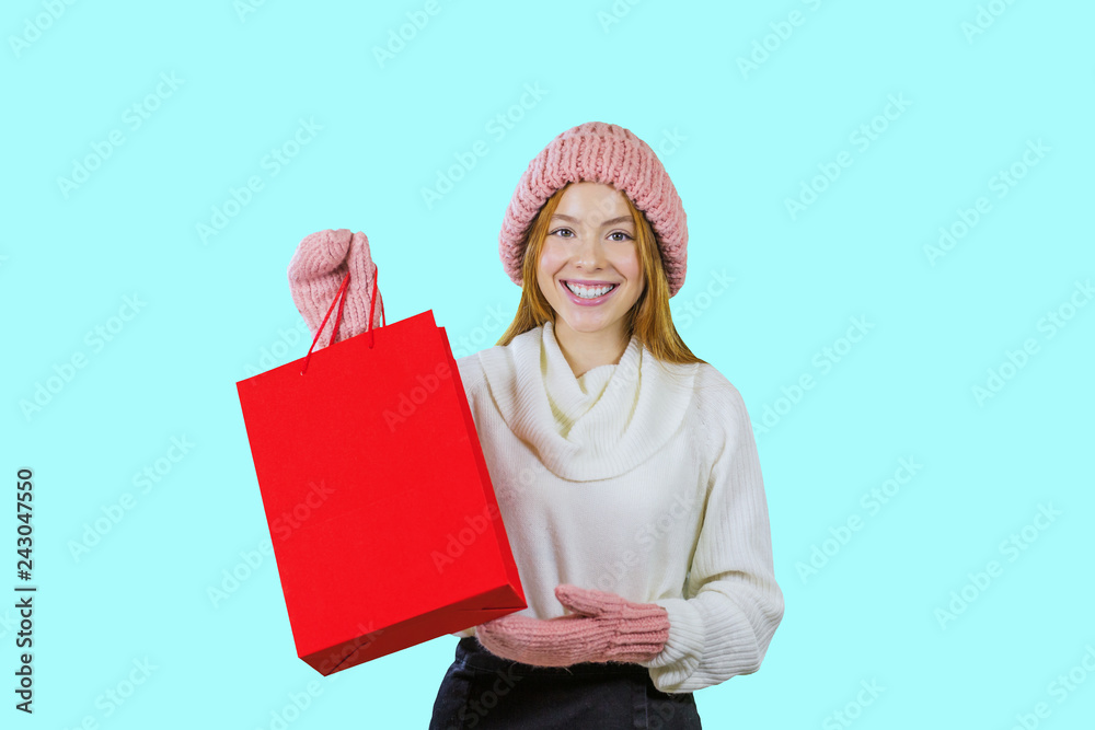 Portrait of a cute red-haired girl in a knitted hat and mittens holding a red bag and looking at the camera smiling brightly showing a gift on an isolated background, fashion shooting, New Year's Portrait of a cute red-haired girl in a knitted hat and mittens holding a red bag and looking at the camera smiling brightly showing a gift on an isolated background, fashion shooting, New Year's