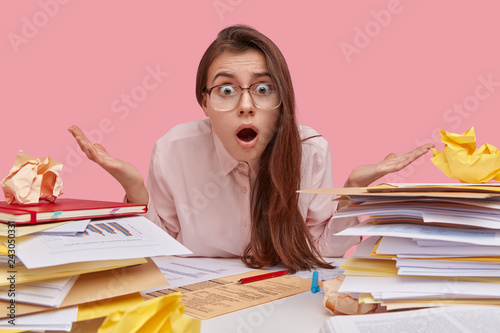 Emotive young European woman with dark hair, feels puzzled and hesitant, opens mouth with bewilderment, wears spectacles and shirt, has stack of papers and books, isolated over pink background