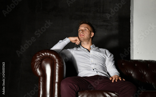 Young business man sitting in leather Chester armchair sofa in casual shirt thinking