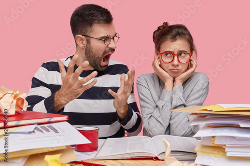 Dissatisfied bearded young male tutor gestures angrily screams at lazy trainee who arent ready with home assignment, sit near opened book and heap of paper documents, study new topic together photo