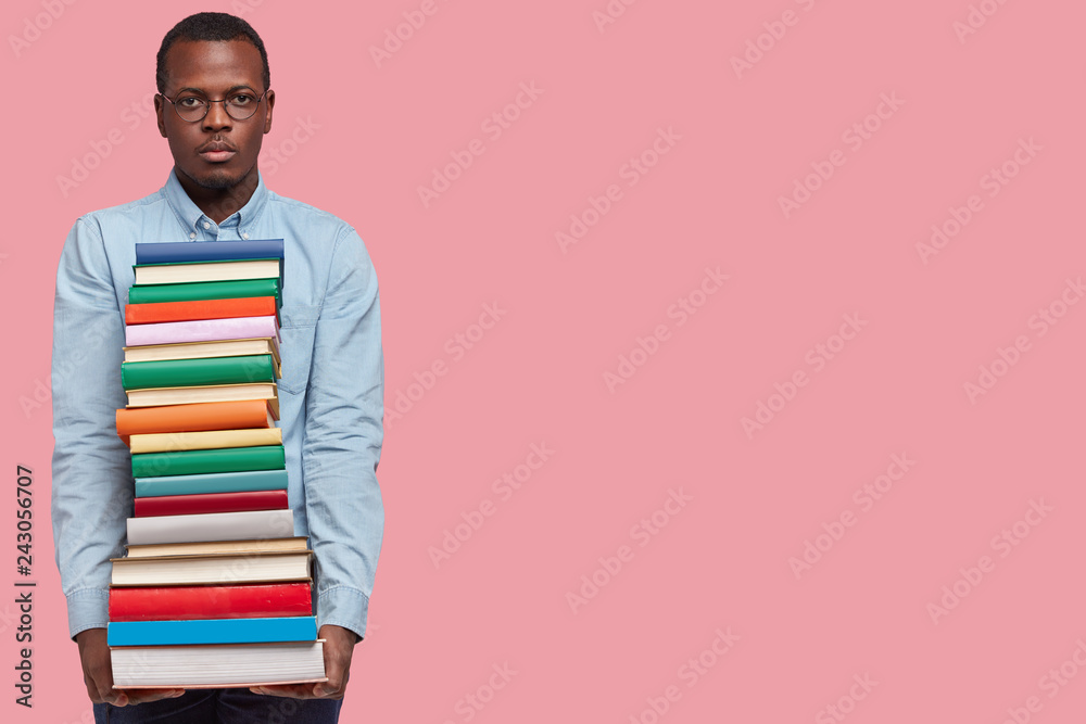 Studio shot of dark skinned serious confident college student carries many books in pile, wears glasses and shirt, returns from library, chooses literature for writing course paper, isolated on pink Studio shot of dark skinned serious confident college student carries many books in pile, wears glasses and shirt, returns from library, chooses literature for writing course paper, isolated on pink