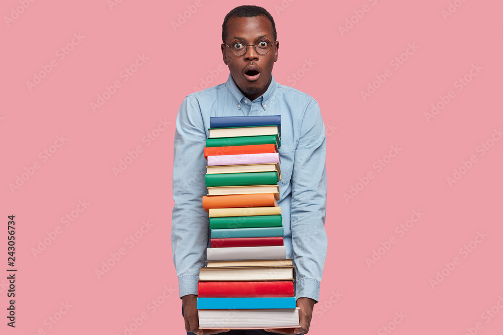 Shot of surprised African American man carries pile of colourful books, scared of having deadline, dressed in formal wear, keeps jaw dropped, isolated over pink background, analyzes literature Shot of surprised African American man carries pile of colourful books, scared of having deadline, dressed in formal wear, keeps jaw dropped, isolated over pink background, analyzes literature