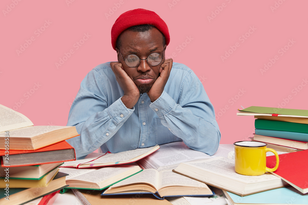 Upset black young man focused down, feels depressed and sad as reads books for long time, wears round spectacles, red hat and shirt, has dejected facial expression, isolated over pink studio wall